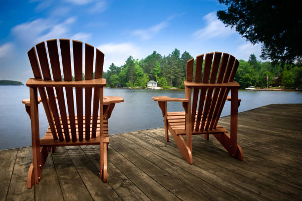 istock Two chairs on a dock face a serene lake, framing a distant white cottage amid lush greenery. 1817664123