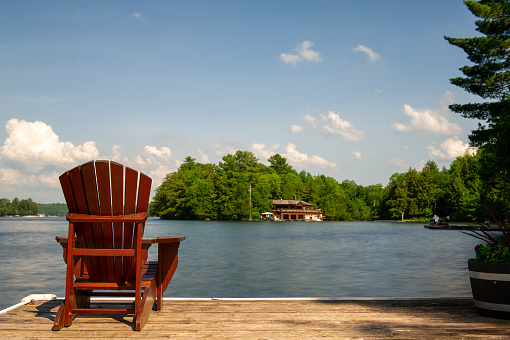 On the wooden dock, a lone Muskoka chair offers a tranquil view of the serene lake. Across the water, a cottage is nestled amidst a lush backdrop of green trees.