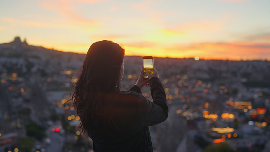 A multiracial female tourist is using her mobile smart phone to take photos and videos of the sunset from top of a hill in Cappadocia Türkiye Turkey.