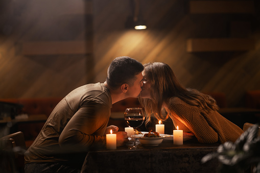 Happy young couple in love kissing, drinking red wine having romantic dinner, celebrating Valentines day, anniversary, sitting at home. Copy space