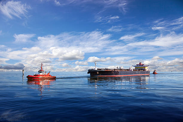 front view on a tugboat towing a large cargo container ship with piled up cargo containers on deck into harbor.