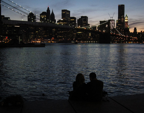 A couple sitting in Main Street Park (part of Brooklyn Bridge Park) enjoy the view of Brooklyn Bridge and the sun setting behind Lower Manhattan in the background.