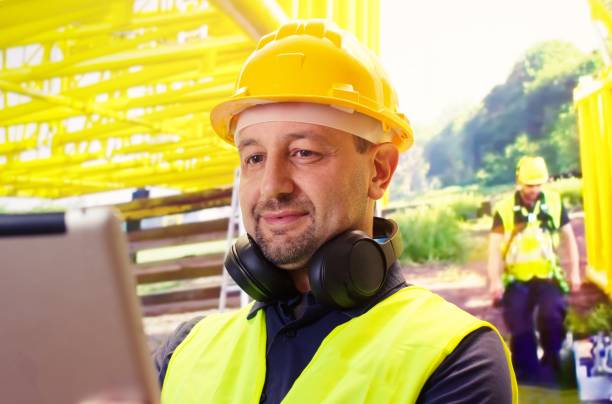 黄色いヘルメットをかぶって微笑む陽気な白人労働者がタブレットを探しています。 - construction worker manual worker construction group of people ストックフォトと画像