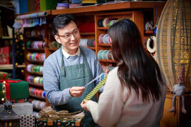 Asian male craftsperson choosing ribbon for customer in christmas shop.