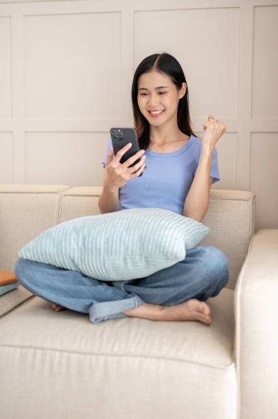 istock Young woman using smartphone and celebrating for good news while sitting on the sofa. 1818038233