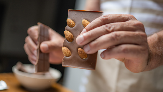 Senior man hands breaking chocolate with almond and putting pieces in bowl close up