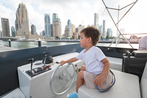 Travel scene in Dubai Marina captures a toddler seated at the yacht's helm, experiencing the joy of navigating amidst the urban splendor and shimmering waters in the United Arab Emirates. 2 year old boy visiting Dubai at taking the cruse ride