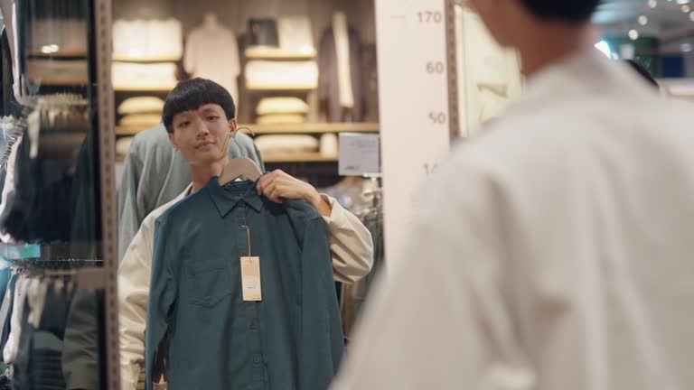 An Asian man holding clothes up to the mirror at the clothing store