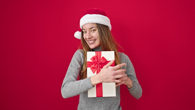 Young caucasian woman wearing christmas hat holding gift over isolated red background
