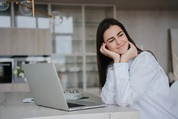 Positive brunette girl in white shirt sitting at desk with laptop leaning on hands looks aside with cute smile on face makes break of remote work at home. Home office, business ideas.
