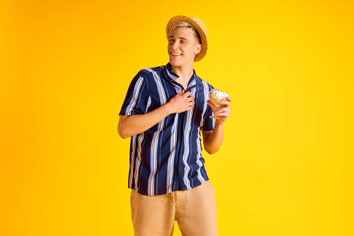 Handsome smiling young man in striped shirt, shorts and straw hat eating delicious vanilla ice cream against yellow studio background. Concept of human emotions, youth, summer vacation, fashion