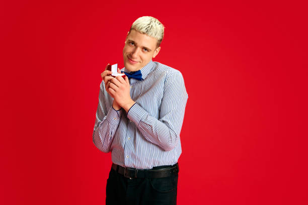Young smiling man in shirt with bow tie, holding ring gift against red studio background. Making proposal. Concept of human emotions, youth, Christmas, winter holidays, vacation, marriage, celebration