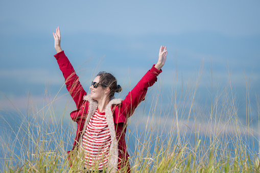 A cheerful young woman in a red jacket stands on top of a green hill, arms spread.