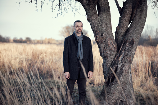 Man in a coat stands near a tree in a park in the countryside. Rest, slowing down, meditative mood.