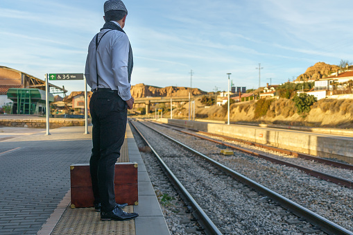 A man alone on the platform next to the train tracks, his suitcase on the ground, looks to his left to see the arrival of the railway.