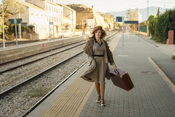 A lady walks on the platform next to the train tracks, she is a tourist in the city and she is wearing a very romantic vintage model.