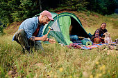 Happy man building a tent while camping with his girlfriend and dog in nature.