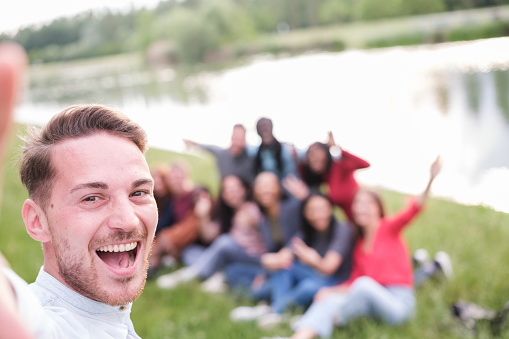 Young man taking a selfie with his group of friends out of focus behind enjoying outdoors