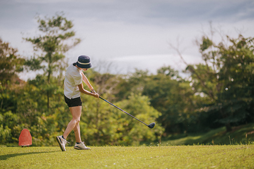 Rear view of asian chinese young female golfer teeing off and swing her driver club on the golf course