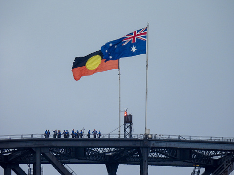 The Australian and Australian Aboriginal flags, appearing faded, weathered and frayed, fly on the Sydney Harbour Bridge.  A group of people have climbed the bridge and looking at the view. This image was taken on an overcast and rainy afternoon on 25 November 2023.