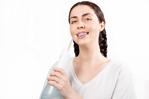 Portrait of young smiling woman with brackets on teeth cleans them using oral irrigator. White background. Concept of dental care during orthodontic treatment.