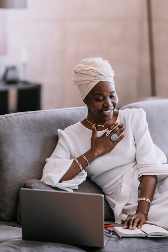 surprised African woman in a turban in a white dress, sitting on the couch at home, working with a laptop from from opened a diary, was surprised at the things done. Productive time management.