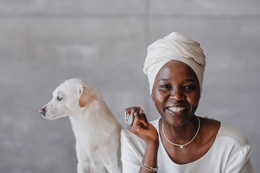 cheerful African woman in traditional clothes with jewelry smiles looks at the camera spends time at home with a pet with a white dog on the background of a wall with a free place