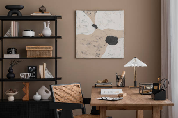 istock Creative composition of warm and cozy office interior with mock up poster frame, wooden desk, rattan chair, stylish lamp, black rack, books and office accessories. Home decor. Template. 1818325749