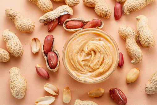 Bowl of peanut butter and peanuts around it on beige background. Top view.