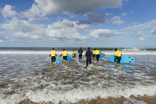 A wide angle rear view of a group of friends who are all taking part in a surfing lesson along the coast in Ambleside in the North East of England. The instructor is telling them to brave the cold and they will climatized to it once in the water.