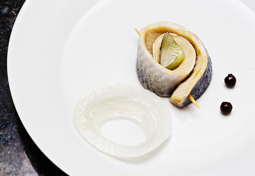 A rollmop, pickled herring fillets rolled around a sliced pickled gherkin. Rollmops are a traditional dish that can be traced back to Northern Europe and have been a staple food. They gained popularity in Germany in the early 19th century and are now commonly eaten in large parts of the world, and are also associated with Ashkenazi Jewish cuisine. Sliced pickled onion and allspice berries, used to flavor the marinade, are also seen in this photo.