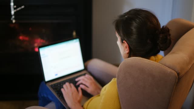 A woman alone at home in front of a fireplace with a laptop chatting, consulting information and social networks, and writing emails during the sunset.
