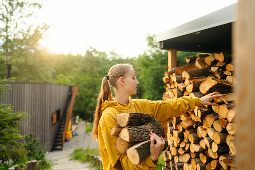 Photo of a young woman bringing the chopped firewood into the house so she can lit a fire. She is trying out a new sustainable lifestyle during her stay in a house rental on a remote location.