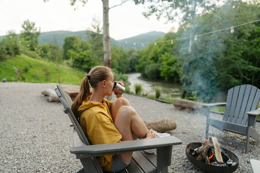 Photo of a young woman relaxing in a garden chair, sipping coffee and overlooking the river from her back yard, as the fire is crackling from the fire pit in front of her