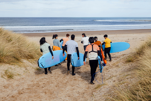 A wide angle rear view of a group of friends who are all taking part in a surfing lesson along the coast in Ambleside in the North East of England. The instructor is telling them to brave the cold and they will climatized to it once in the water. They are walking through a sandy area heading to the shore.