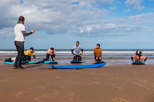 A wide angle view of a surfing class on the beach in Ambleside. The group of friends are lined up and the instructor is going over safety before they all go in the water together. He is talking about kneeling on the board and also showing them the surf straps.