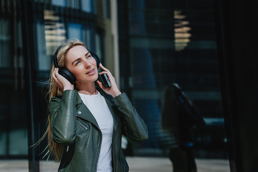 Dreaming hipster blonde girl in leather jacket listening music outside uses headphones walking by street relaxing on sunny summer day, traveling explores new city. Mockup, leisure, carefree people.