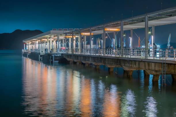 Hong Kong, Hong Kong – November 11, 2021: A scenic nighttime view of a dock illuminated by bright lights, situated in front of the majestic ocean and mountain ranges in the background