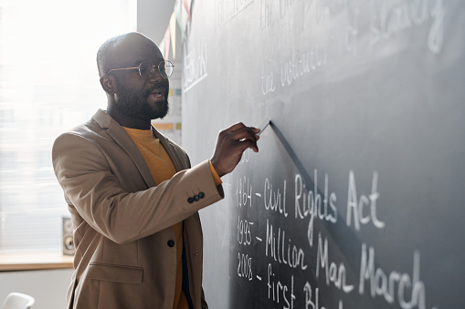 African American teacher writing information on blackboard with chalk during lesson at school
