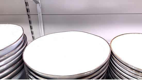 New modern plates of various shapes and shades for eating are stacked in a row on the shelf of a tableware store. A group of ceramic plates stacked on top of each other on a shelf. Crockery, china, tableware