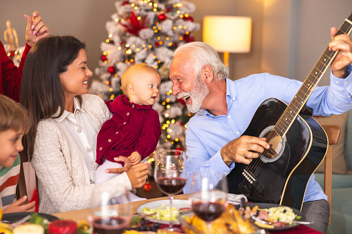 Happy multi-generation family gathered around the table playing the guitar and singing Christmas songs while having Christmas dinner all together at home