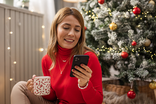Young woman celebrating Christmas at home. She is decorating Christmas tree, enjoying in coffee and cookies or using smart phone.