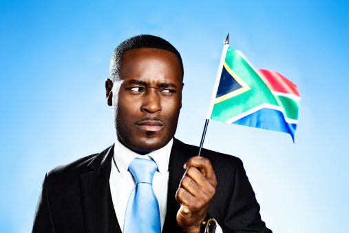 A frowning  African man holds up the distinctive South African national flag against a sky-blue background. He is obviously not a happy citizen!