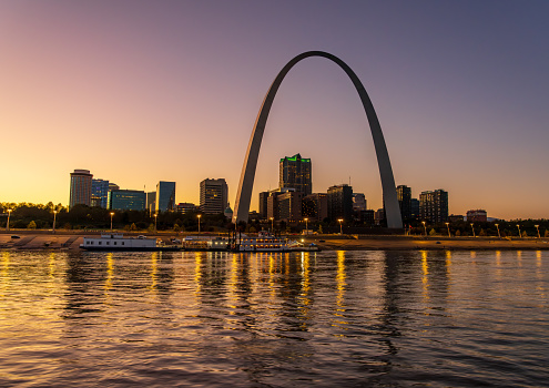 The city of St. Louis, Missouri, as seen from across the river.  The Gateway Arch dominates the left upper section of the photo, more than twice as high as any of the buildings in the photo.  In a horizontal line across the background and behind the Gateway Arch are downtown buildings and some skyscrapers.  Lights are just coming on in the buildings even though there is still light in the sky.  A line of beach lies between the buildings and the river, which is a dark blue, mirroring the dark blue of the sky.