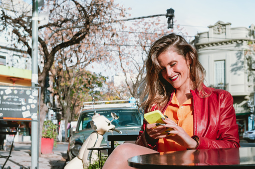 pretty young hispanic latina blonde girl in red jacket and orange shirt sitting outdoors happy and smiling checking her phone, lifestyle concept.
