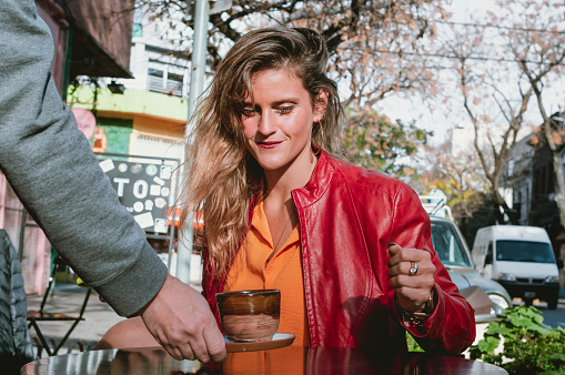 beautiful young hispanic latina blonde girl with red jacket and orange shirt sitting outdoors happy and smiling watching a cup of coffee being served on the table. lifestyle concept.