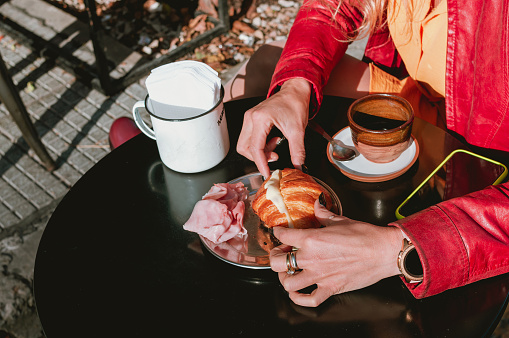 Close up shot of a cup of coffee, a telephone and the pretty feminine hands of a blonde girl, holding a ham and cheese croissant on a stainless steel plate, on a black outdoor table.lifestyle concept.