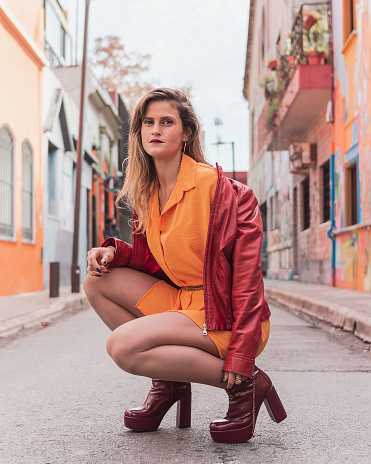 portrait of young beautiful Hispanic Latin blonde girl with red jacket, orange dress and nice legs, posing crouching in the middle of a street with colorful walls.