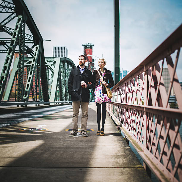 A smiling well dressed young couple stand on the Hawthorne bridge over the Willamette river in Portland, Oregon.  Square crop with copy space, the city visible in the background.