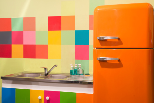 Vibrant colorful kitchen with bright orange refrigerator and rainbow checkerboard wall and cabinets surrounding the sink.